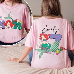May include: Pink t-shirt featuring a mermaid design. The front of the shirt has the word "MOM" with a mermaid illustration. The back of the shirt has the name "Emily" and a mermaid illustration with the number "7".