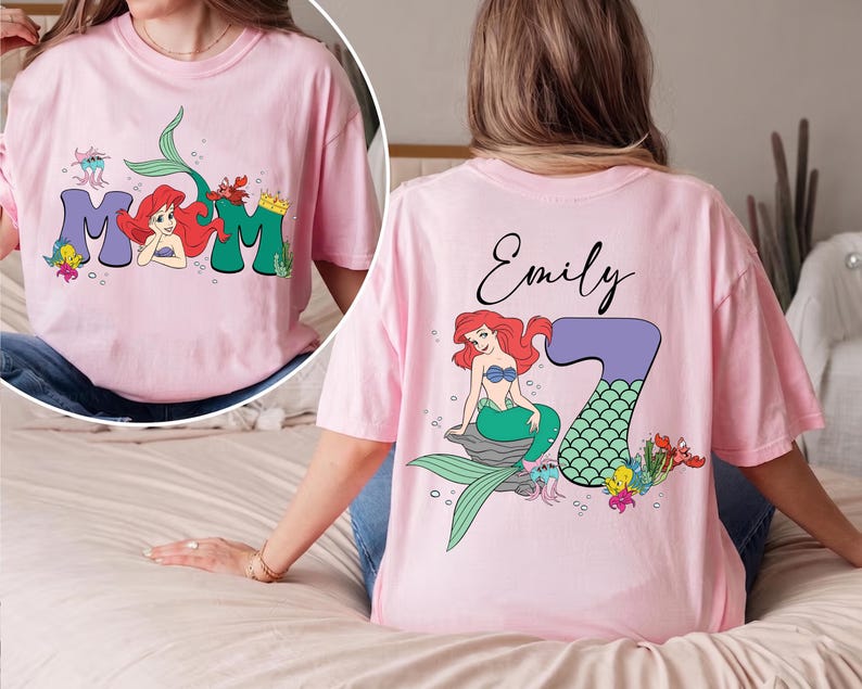 May include: Pink t-shirt featuring a mermaid design. The front of the shirt has the word "MOM" with a mermaid illustration. The back of the shirt has the name "Emily" and a mermaid illustration with the number "7".
