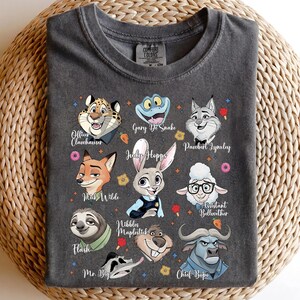 May include: A charcoal gray t-shirt featuring a graphic print of various animated animal characters. The characters include a cheetah, snake, lynx, fox, rabbit, sheep, sloth, beaver, and buffalo, each with their name printed in white script.