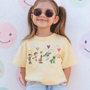 Op de afbeelding: Lichtgeel T-shirt met een cartoonafbeelding van Toy Story-personages die ballonnen vasthouden. Het ontwerp omvat Woody, Jessie, Buzz Lightyear en andere personages. Het T-shirt wordt gedragen door een jong meisje met een ronde zonnebril.