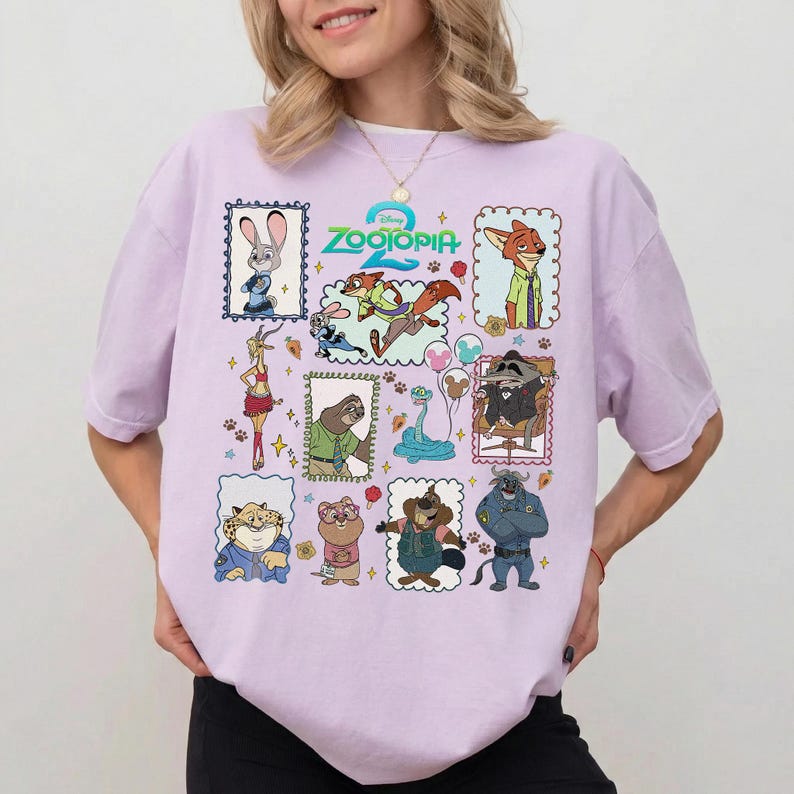 May include: A light purple short-sleeve t-shirt featuring a colorful graphic print of various Zootopia characters in individual frames, including Judy Hopps, Nick Wilde, Flash, and others, with the Zootopia 2 logo centered at the top of the design.