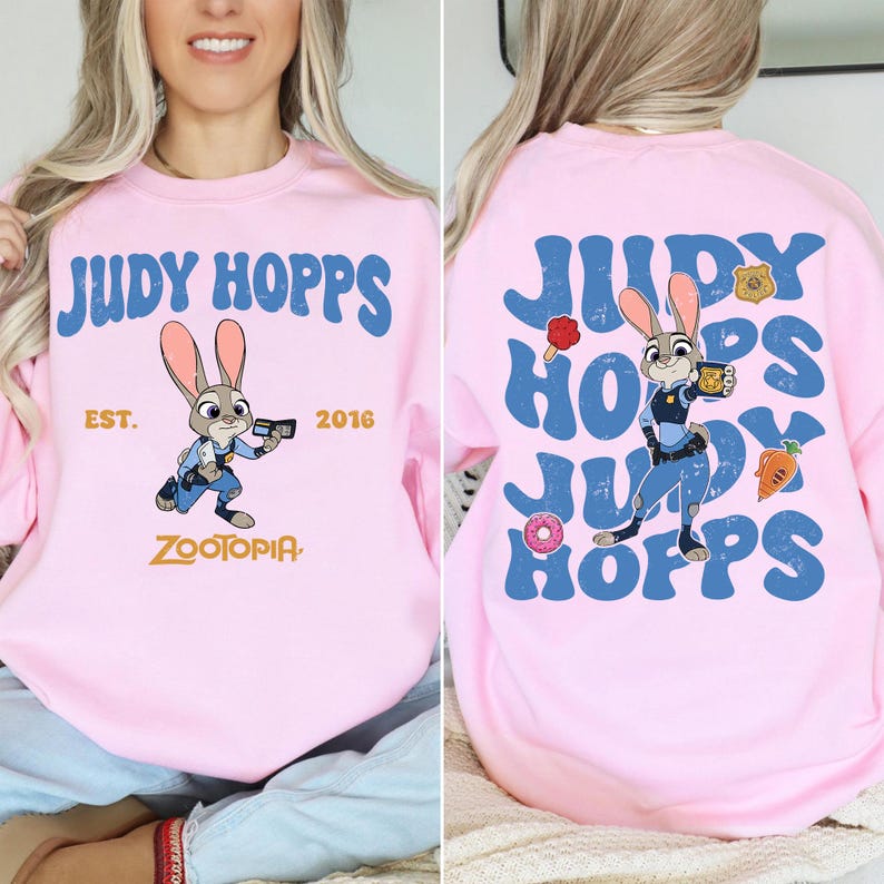 May include: A light pink sweatshirt featuring Judy Hopps from Zootopia. The front displays the text Judy Hopps, Est. 2016, and Zootopia with a graphic of the character. The back shows a large repeating Judy Hopps text with the character and icons.
