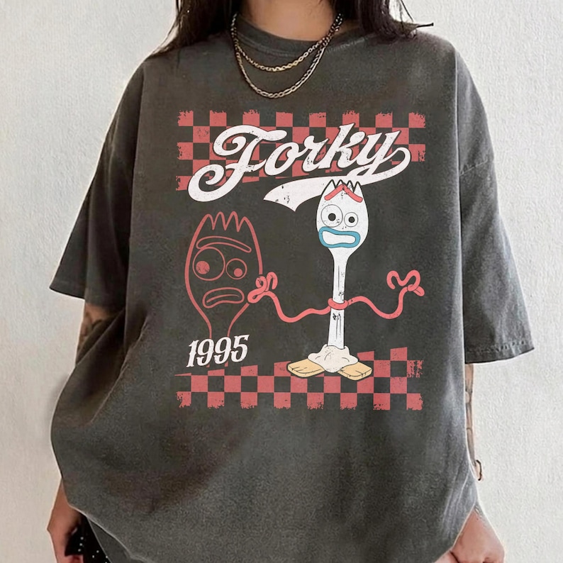May include: Dark gray t-shirt featuring the word "Forky" above a cartoon fork character with a red and white checkered background. The year "1995" is printed below a cartoon fork. The shirt has short sleeves.