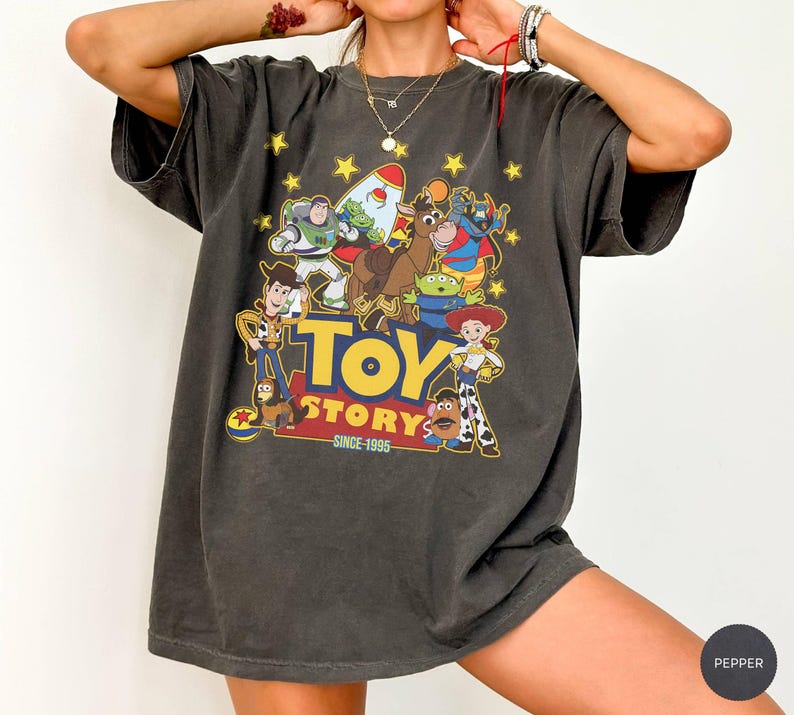 May include: Dark gray t-shirt featuring a colorful graphic of Toy Story characters, including Woody, Buzz Lightyear, and Jessie. The text "TOY STORY" is in yellow and blue, with "SINCE 1995" below. The shirt has a vintage, worn look.