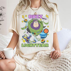 May include: Cream-colored t-shirt featuring a graphic of Buzz Lightyear from Toy Story. The design includes the text "BUZZ LIGHTYEAR" in purple and green, with stars and planets. The shirt is short-sleeved.