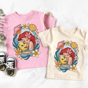 May include: Two short-sleeved t-shirts, one pink and one cream, featuring a cartoon graphic of a red-haired mermaid hugging a yellow fish. The design is framed by seashells, flowers, and a blue border. Black and white sneakers are visible.