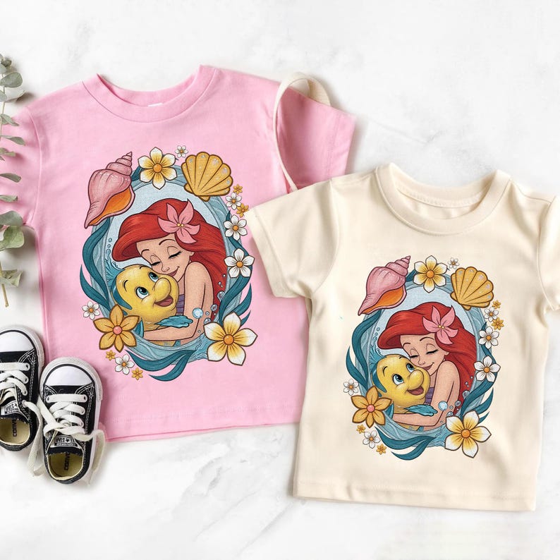 May include: Two short-sleeved t-shirts, one pink and one cream, featuring a cartoon graphic of a red-haired mermaid hugging a yellow fish. The design is framed by seashells, flowers, and a blue border. Black and white sneakers are visible.