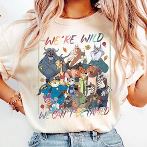 May include: An ivory t-shirt featuring a colorful graphic print of various animated animal characters. The text "WE'RE WILD" appears at the top and "WE CAN'T BE TAMED" at the bottom in multi-colored lettering. The design includes a variety of creatures.