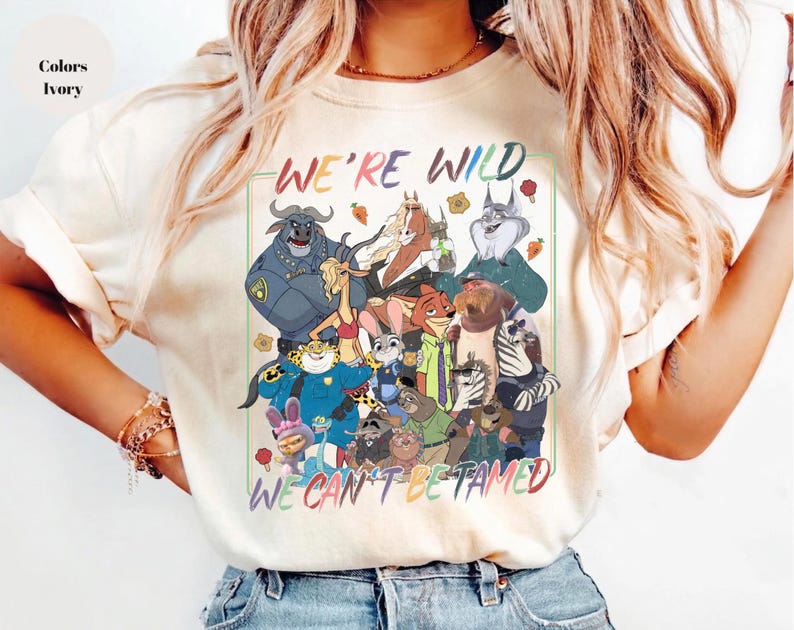 May include: An ivory t-shirt featuring a colorful graphic print of various animated animal characters. The text "WE'RE WILD" appears at the top and "WE CAN'T BE TAMED" at the bottom in multi-colored lettering. The design includes a variety of creatures.