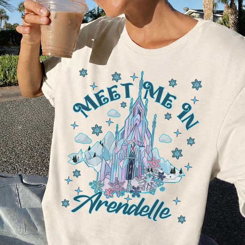 May include: Cream-colored t-shirt featuring the text "Meet Me In Arendelle" in teal script. The graphic includes a stylized ice castle, mountains, and snowflakes in shades of blue, purple, and pink. A casual, comfortable tee.