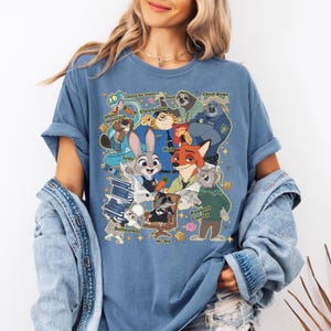 Op de afbeelding: Blauw T-shirt met een kleurrijke afbeelding van personages uit de animatiefilm Zootopia. Het ontwerp bevat Judy Hopps, Nick Wilde en andere personages met hun namen. Het shirt heeft opgerolde mouwen.