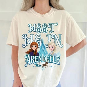 May include: Cream-colored t-shirt featuring the text "Meet Me In Arendelle" in teal script. The design includes illustrations of two animated female characters, a castle, and a reindeer. The shirt is a short-sleeved crew neck.