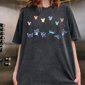 May include: A charcoal gray short-sleeve t-shirt featuring a graphic print of the character Stitch in various poses, each holding a string attached to a pastel-colored balloon shaped like a mouse head, accented with small white star illustrations.