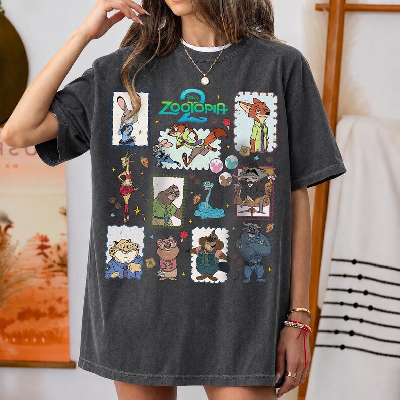 May include: A charcoal gray short-sleeve t-shirt featuring a collage of various Zootopia characters in rectangular frames. The graphic includes the Zootopia logo in teal at the top, with illustrations of Judy Hopps, Nick Wilde, Flash, and other movie characters.