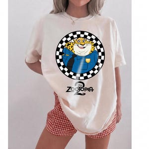 May include: An off-white t-shirt featuring a graphic of the Zootopia character Clawhauser in a blue police uniform, enclosed within a black and white checkered circle. The text Zootopia 2 is printed in black below the circular graphic design.