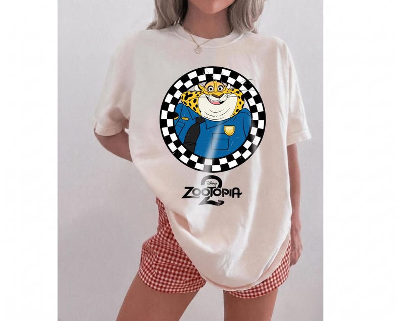 May include: An off-white t-shirt featuring a graphic of the Zootopia character Clawhauser in a blue police uniform, enclosed within a black and white checkered circle. The text Zootopia 2 is printed in black below the circular graphic design.