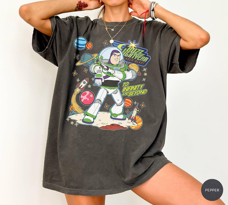 May include: Dark gray t-shirt featuring a graphic of Buzz Lightyear from Toy Story. The design includes the text "Buzz Lightyear Laser Blast" and "To Infinity and Beyond" with planets, rockets, and stars.