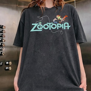 May include: A charcoal gray short-sleeve t-shirt featuring the light blue text Zootopia across the chest. The graphic includes a small green snake, a white rabbit, and an orange fox character positioned above the lettering with a dashed line.