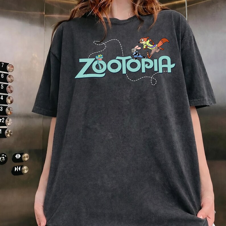 May include: A charcoal gray short-sleeve t-shirt featuring the light blue text Zootopia across the chest. The graphic includes a small green snake, a white rabbit, and an orange fox character positioned above the lettering with a dashed line.