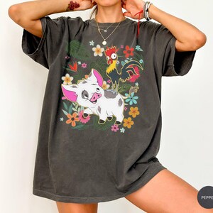 Puede incluir: Camiseta gris oscuro con un dise&ntilde;o de cerdo y gallo de dibujos animados rodeados de flores y follaje coloridos. El dise&ntilde;o gr&aacute;fico incluye elementos florales rosas, amarillos y azules. La camiseta tiene mangas cortas y un corte holgado.