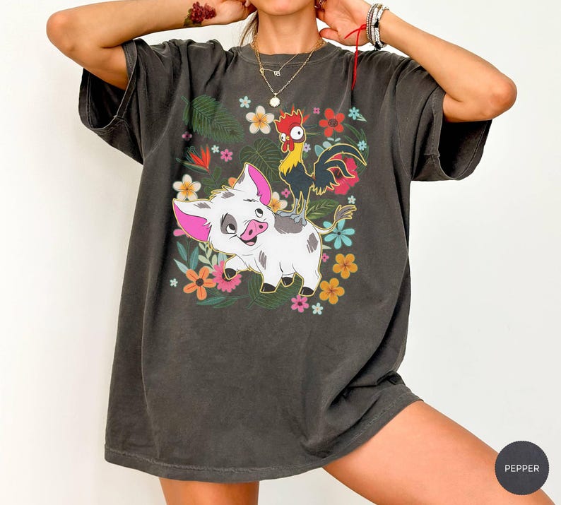 Puede incluir: Camiseta gris oscuro con un dise&ntilde;o de cerdo y gallo de dibujos animados rodeados de flores y follaje coloridos. El dise&ntilde;o gr&aacute;fico incluye elementos florales rosas, amarillos y azules. La camiseta tiene mangas cortas y un corte holgado.