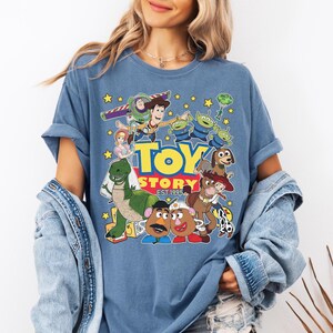 May include: Blue t-shirt featuring a colorful graphic of Toy Story characters, including Woody, Buzz Lightyear, and the Little Green Men. The text "TOY STORY EST. 1995" is also displayed. The shirt has short sleeves and a relaxed fit.