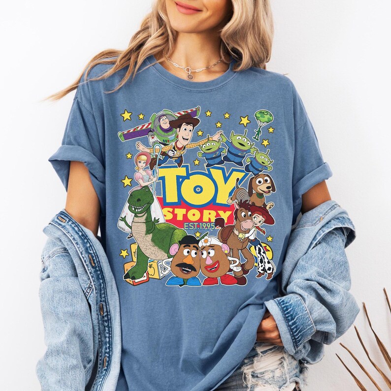 May include: Blue t-shirt featuring a colorful graphic of Toy Story characters, including Woody, Buzz Lightyear, and the Little Green Men. The text "TOY STORY EST. 1995" is also displayed. The shirt has short sleeves and a relaxed fit.