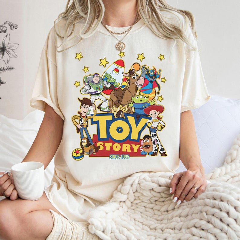 May include: Cream-colored t-shirt featuring a colorful graphic of Toy Story characters, including Woody, Buzz Lightyear, and others, with the words "TOY STORY" and "SINCE 1995". The design is surrounded by yellow stars.