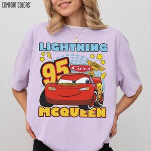 May include: A light purple Comfort Colors t-shirt featuring a graphic of Lightning McQueen from the Cars movie. The design includes the number 95, the word "Lightning" above the car, and the word "McQueen" below. The shirt has a crew neck.