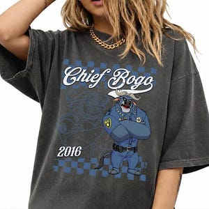 Puede incluir: Una camiseta gris carb&oacute;n de corte oversize con un estampado gr&aacute;fico de un b&uacute;falo polic&iacute;a de dibujos animados con uniforme azul y brazos cruzados. El texto "Chief Bogo" aparece en letra blanca sobre el personaje, con el a&ntilde;o "2016" debajo.