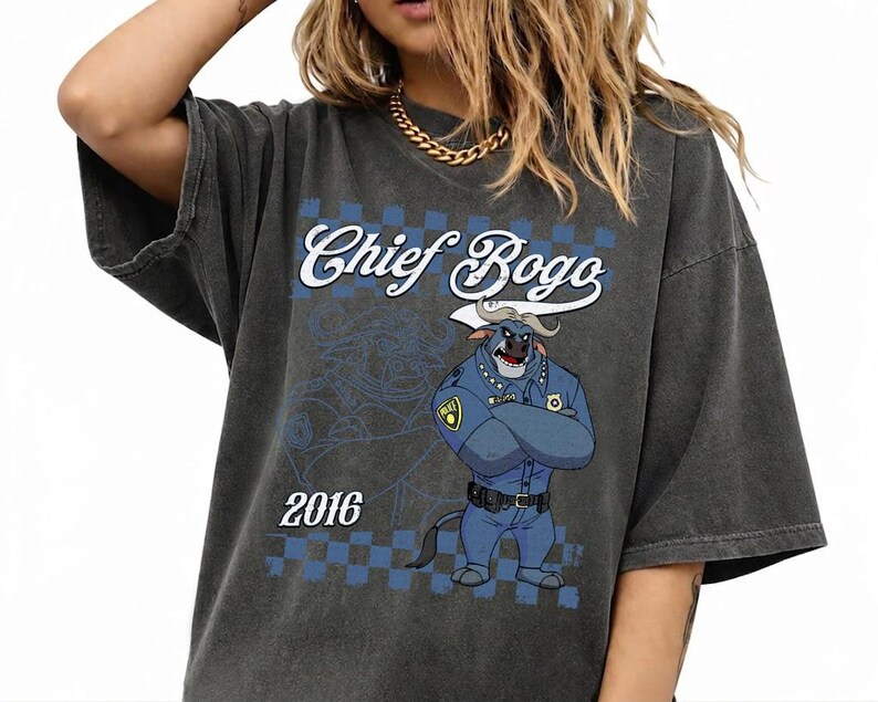 Puede incluir: Una camiseta gris carb&oacute;n de corte oversize con un estampado gr&aacute;fico de un b&uacute;falo polic&iacute;a de dibujos animados con uniforme azul y brazos cruzados. El texto "Chief Bogo" aparece en letra blanca sobre el personaje, con el a&ntilde;o "2016" debajo.