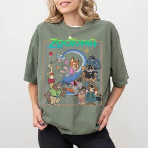 May include: An olive green short-sleeve t-shirt featuring a graphic print of Zootopia characters including Nick Wilde, Judy Hopps, Flash, and others. The design is enclosed in a rectangular orange border with the Zootopia logo at the top.