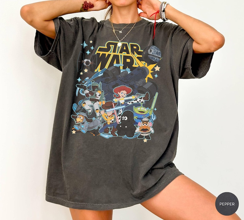 May include: Dark gray t-shirt featuring a graphic of Star Wars characters and Toy Story characters. The Star Wars logo is in yellow and the shirt has a vintage, worn look.