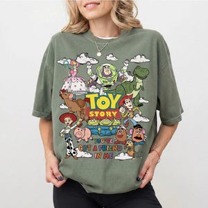 Op de afbeelding: Olijfgroen T-shirt met een kleurrijke afbeelding van personages uit de animatiefilm "Toy Story". Het ontwerp bevat Woody, Buzz Lightyear, Rex en andere personages, met de tekst "Toy Story" en "You've Got a Friend in Me."