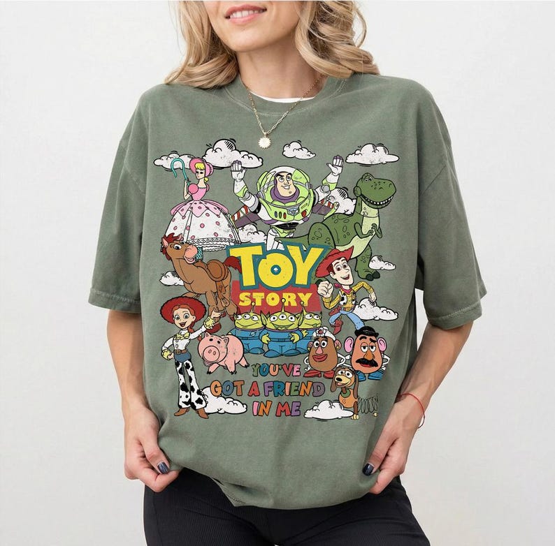 Op de afbeelding: Olijfgroen T-shirt met een kleurrijke afbeelding van personages uit de animatiefilm "Toy Story". Het ontwerp bevat Woody, Buzz Lightyear, Rex en andere personages, met de tekst "Toy Story" en "You've Got a Friend in Me."