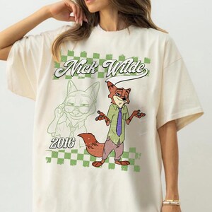 Puede incluir: Una camiseta de color crema con un gr&aacute;fico del personaje Nick Wilde. El dise&ntilde;o incluye un fondo de cuadros verdes y blancos, el texto "Nick Wilde" en letra cursiva, el a&ntilde;o "2016" y una ilustraci&oacute;n a todo color del personaje zorro.