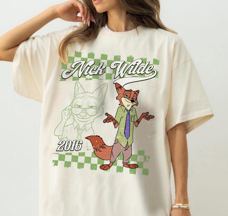Puede incluir: Una camiseta de color crema con un gr&aacute;fico del personaje Nick Wilde. El dise&ntilde;o incluye un fondo de cuadros verdes y blancos, el texto "Nick Wilde" en letra cursiva, el a&ntilde;o "2016" y una ilustraci&oacute;n a todo color del personaje zorro.