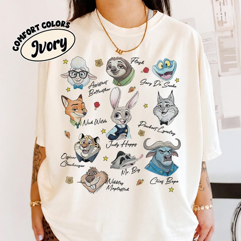 May include: An ivory t-shirt featuring a graphic print of various animated animal characters from Zootopia, including Judy Hopps, Nick Wilde, and Chief Bogo, with their names printed in black script alongside small star and carrot icons.