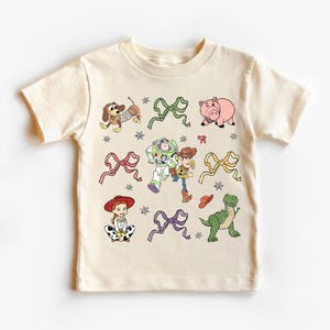 Op de afbeelding: Cr&egrave;mekleurig T-shirt met kleurrijke cartoonfiguren en strikken. Het ontwerp bevat een teckel, een roze varken, Buzz Lightyear, Woody, Jessie en een groene dinosaurus. Het shirt heeft een ronde hals en korte mouwen.