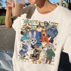Op de afbeelding: Cr&egrave;mekleurig T-shirt met een kleurrijke cartoonillustratie van verschillende dierenfiguren. Het ontwerp bevat een slang, een luiaard, een vos, een konijn en andere personages. Tekst bevat karakternamen zoals "Judy" en "Nick".