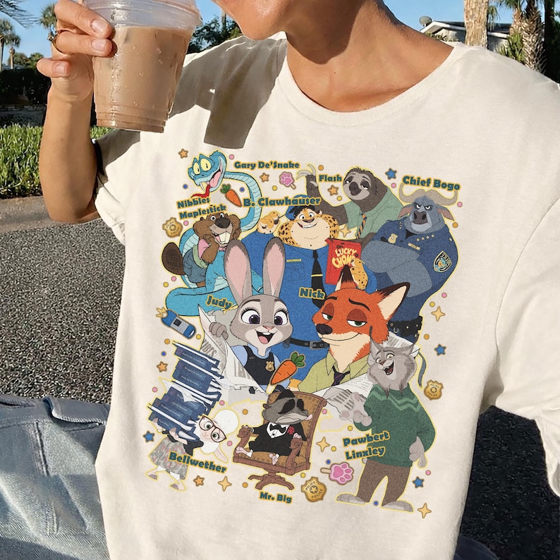 Op de afbeelding: Cr&egrave;mekleurig T-shirt met een kleurrijke cartoonillustratie van verschillende dierenfiguren. Het ontwerp bevat een slang, een luiaard, een vos, een konijn en andere personages. Tekst bevat karakternamen zoals "Judy" en "Nick".