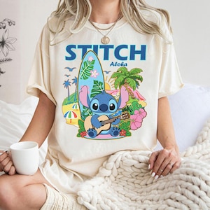 May include: Cream-colored t-shirt featuring a cartoon illustration of Stitch playing a ukulele on a tropical beach. The design includes a surfboard, palm trees, and the word "STITCH" in blue. The word "Aloha" is also included.