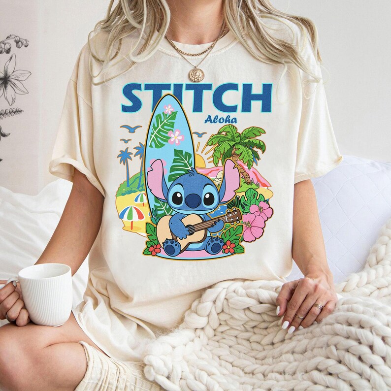 May include: Cream-colored t-shirt featuring a cartoon illustration of Stitch playing a ukulele on a tropical beach. The design includes a surfboard, palm trees, and the word "STITCH" in blue. The word "Aloha" is also included.