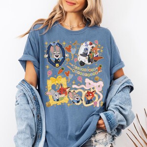 May include: Blue t-shirt featuring cartoon characters from Zootopia. The design includes Judy Hopps, Nick Wilde, and other characters in frames and a police vehicle. The text "WELCOME TO ZOOOTOPIA" is also present.
