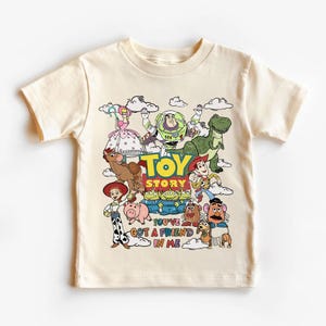 Op de afbeelding: Cr&egrave;mekleurig T-shirt met een kleurrijke afbeelding van personages uit de animatiefilm "Toy Story". Het ontwerp bevat Woody, Buzz Lightyear, Rex en andere personages, met de tekst "TOY STORY" en "YOU'VE GOT A FRIEND IN ME."