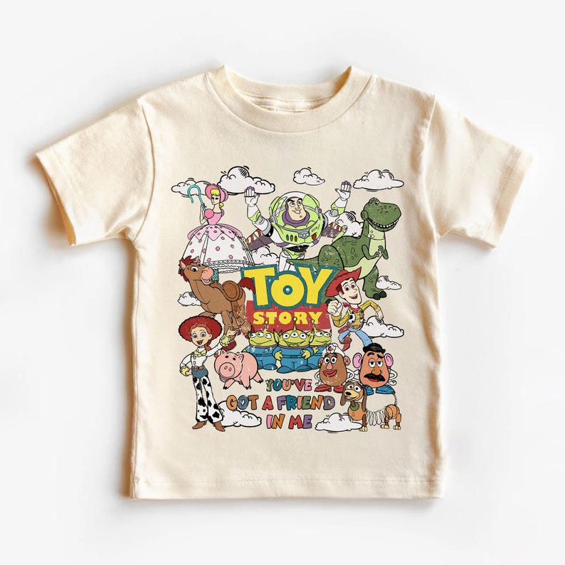 Op de afbeelding: Cr&egrave;mekleurig T-shirt met een kleurrijke afbeelding van personages uit de animatiefilm "Toy Story". Het ontwerp bevat Woody, Buzz Lightyear, Rex en andere personages, met de tekst "TOY STORY" en "YOU'VE GOT A FRIEND IN ME."