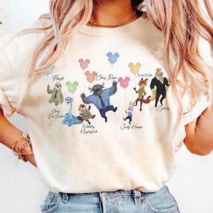May include: An ivory t-shirt featuring a graphic print of various animated characters, including a sloth, snake, beaver, buffalo, rabbit, fox, and horse, each holding a colourful Mickey Mouse-shaped balloon. The names Flash, Gary De Sanka, Nibbles Maplestick, Chief Bogo, Judy Hopps, Nick Wilde, and Zobros are printed below the characters.
