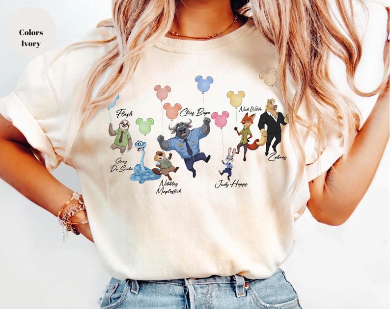 May include: An ivory t-shirt featuring a graphic print of various animated characters, including a sloth, snake, beaver, buffalo, rabbit, fox, and horse, each holding a colourful Mickey Mouse-shaped balloon. The names Flash, Gary De Sanka, Nibbles Maplestick, Chief Bogo, Judy Hopps, Nick Wilde, and Zobros are printed below the characters.