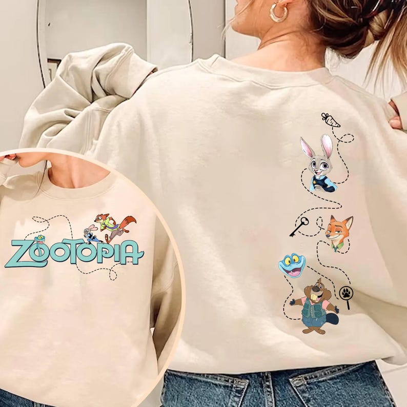 May include: A beige crewneck sweatshirt featuring Zootopia characters. The front displays the Zootopia logo with Judy Hopps and Nick Wilde. The back shows a vertical design with Judy, Nick, Flash, and Mr. Big connected by a dotted line with icons.
