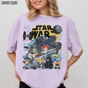 May include: A light purple Comfort Colors t-shirt featuring a Star Wars and Toy Story mashup graphic. The design includes characters like Woody, Buzz Lightyear, and Darth Vader, along with the Star Wars logo and iconic elements.